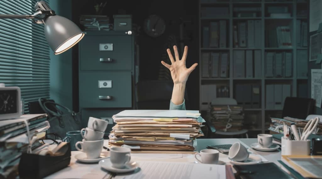 Overwhelmed office worker’s hand reaching from behind piles of paperwork, illustrating AI for SEO digital marketing admin burden.
