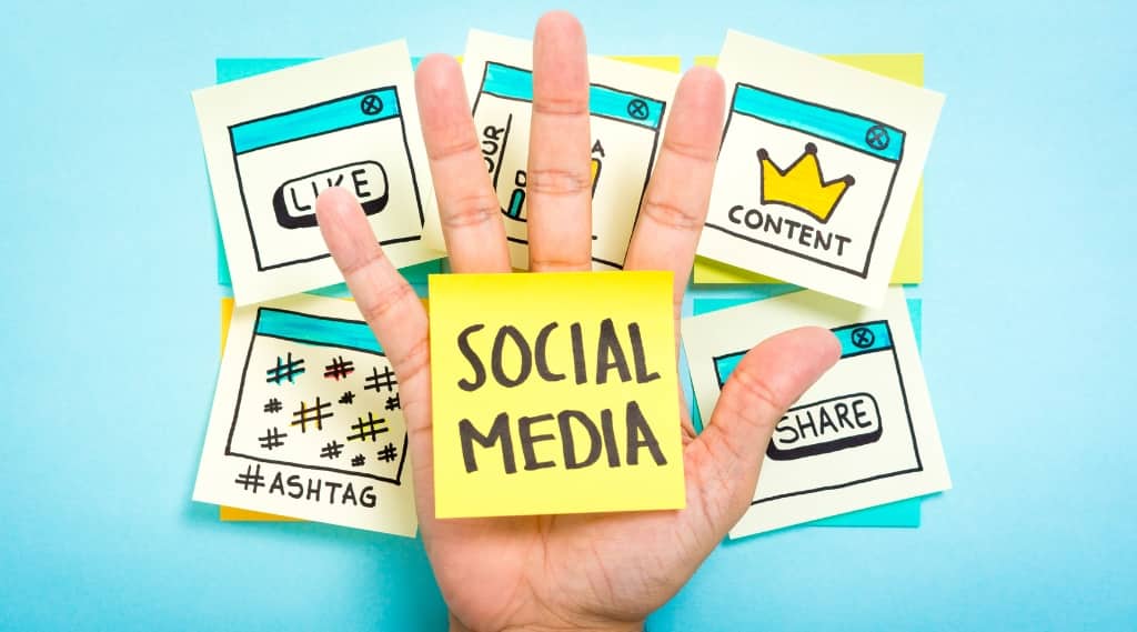 Hand holding sticky note with “Social Media” over background notes for likes, content, and hashtags, representing social media marketing planning.